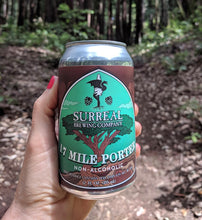 Load image into Gallery viewer, Can of Surreal Non-Alcoholic 17 Mile Porter being held in a hand in the forest setting.

