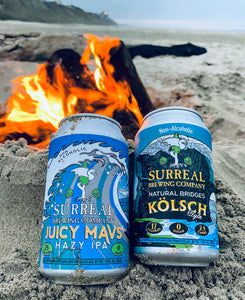 Can of Surreal Non-Alcoholic Juicy Mavs Hazy IPA. 25 calories, zero sugar. with can of Surreal Natural Bridges Kolsch Style. 17 calories. zero sugar. 2.8 carbs. Background fire pit on beach. 