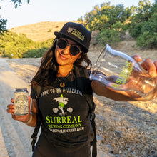 Load image into Gallery viewer, Woman wearing Surreal logo t-shirt and hat holding can of Non-Alcoholic Creatives West Coast IPA and Surreal Branded nucleated glass. 
