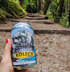 Can of Surreal Non-Alcoholic Kolsch Style. 17 Calories, Zero Sugar, 2.8 calories. Background trail in forest.