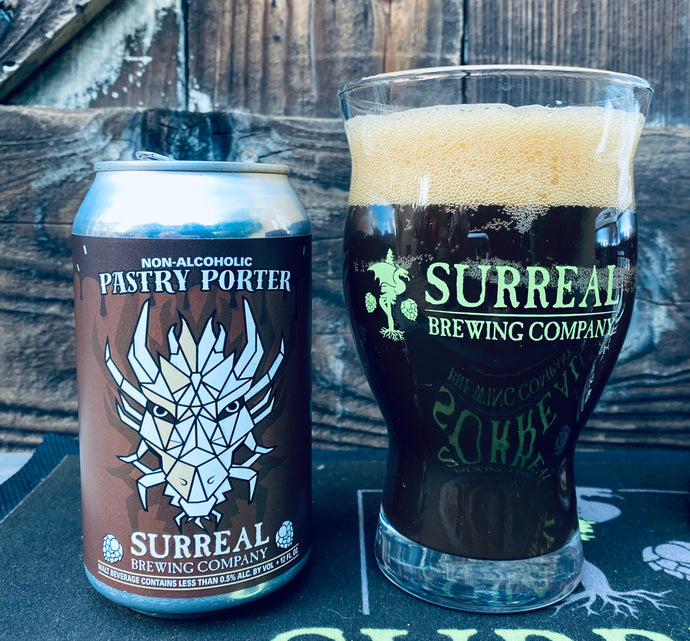 Showing Surreal Branded Nucleated glass with NA Pastry Porter dark colored beer poured out and a thick creamy foamy head. Next to a can of Surreal Non-Alcoholic Pastry Porter
